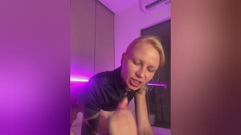 Video of a blonde woman in a black shirt, with a microphone in hand, looking down at a nude man lying on a bed, illuminated by purple lights in a dimly lit room.