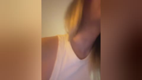 A blurry video of a woman with light skin, wearing a white tank top, her long blonde hair partially visible. The background is out of focus, creating a soft, warm ambiance.