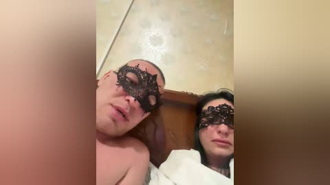 Video of two people in a bathroom, both wearing black, intricate masquerade masks; one person is topless, and the other is partially covered with a white towel.