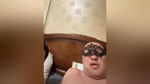 Video of a pale, overweight man with a black masquerade mask, lying on a bed with a beige, floral-patterned lampshade and wooden headboard.