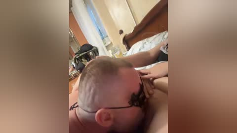 A blurry video shows a man with a bald head and beard, lying on a bed, performing oral sex on a woman with long hair. The room has a wooden headboard, a dresser, and a window with white curtains.