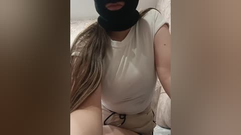 Video of a woman with light skin, wearing a black face mask, white t-shirt, and beige shorts, sitting on a bed with beige sheets, in a dimly lit room.