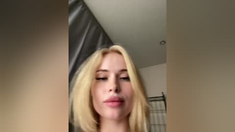 Video of a blonde woman with fair skin, wearing minimal makeup, standing in a bathroom with beige walls and a shower curtain. Her expression is neutral, and the image is slightly blurred.
