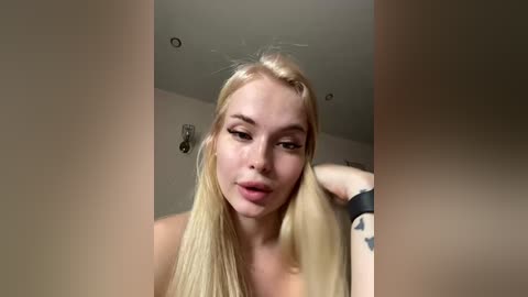 Video of a blonde, light-skinned woman with straight hair, wearing a black wristband, blowing a kiss, in a dimly-lit room with recessed ceiling lights.