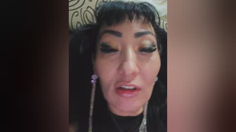 Video of an Asian woman with short black hair and heavy makeup, wearing a black top, against a beige patterned wall.