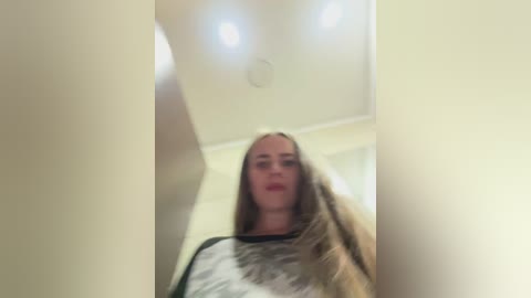 Video of a young woman with long blonde hair, fair skin, and light makeup, wearing a gray and white patterned shirt, standing in a bright, white-walled room with recessed ceiling lights.