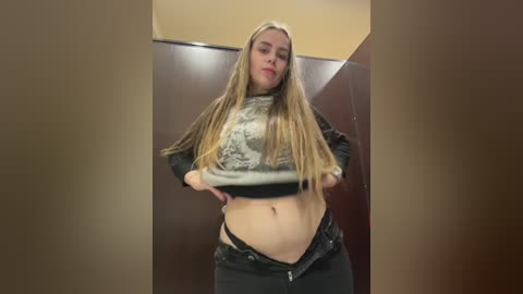 Video of a blonde woman lifting her crop top to reveal her midriff, wearing black jeans, standing in a bathroom stall with beige walls.