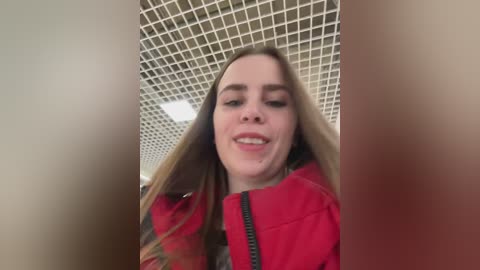 A video of a blonde woman with fair skin, wearing a red jacket, smiling and looking up. She is in an indoor setting with a grid-patterned ceiling and blurred background.