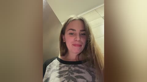 Video of a young Caucasian woman with long, blonde hair, wearing a grey and black camo-print t-shirt. She stands in a narrow hallway with beige walls and tiled floor.