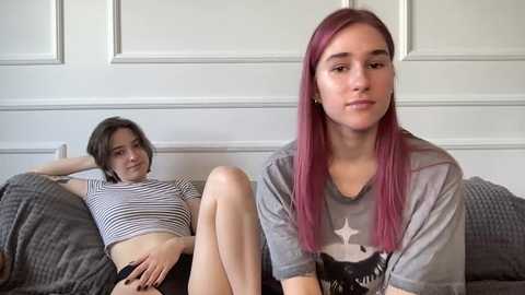 Video of two young women in casual attire, one with short dark hair and the other with long pink hair, lounging on a grey couch in a white-paneled room.