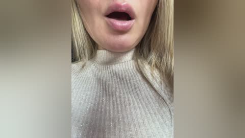 Video of a light-skinned woman with long, straight blonde hair, wearing a beige, ribbed turtleneck sweater. Her mouth is slightly open, revealing slightly parted lips. Background is blurred, focusing attention on her face and sweater.