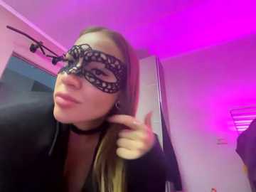 A video of a woman with light skin, wearing a black lace masquerade mask, black choker, and long straight blonde hair, giving a thumbs-up gesture in a room with purple and pink lighting.