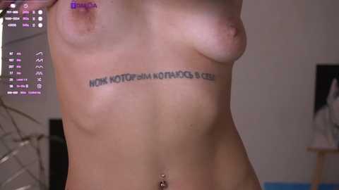 Video of a topless, slender, light-skinned woman with small breasts, displaying a tattoo with Cyrillic text on her ribcage. Background shows a dimly lit room with a TV displaying a blue screen.