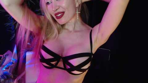 Video of a smiling, light-skinned woman with blonde hair wearing black strappy lingerie and gold hoop earrings, set against a dimly lit, dark background with colorful lights.