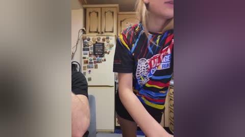 Video of a young woman with blonde hair, wearing a colorful striped shirt, and shorts, standing in a cluttered kitchen with a refrigerator adorned with magnets.