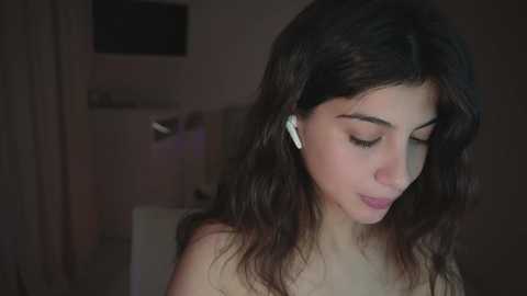 Video of a young woman with long dark hair, wearing white earbuds, and looking down, possibly listening to music, in a dimly lit room with a TV and blurred background.