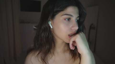 A close-up video of a young woman with long dark hair, wearing white earphones, biting her finger, with a pensive expression, in a dimly lit room.