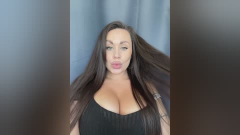 Video of a light-skinned woman with long, dark brown hair, wearing a low-cut black dress, puckering her lips, against a blurred gray background.