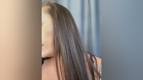 A video of a woman with long, straight brown hair, partially obscured by a vertical light-colored curtain, standing against a neutral blue-gray wall. She wears a black top, her face is not visible.