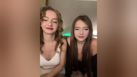 Video of two young women, one with curly hair, the other with straight hair, smiling, wearing white tops, in a dimly lit room with green lights.