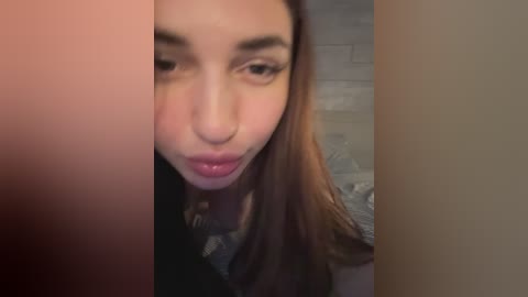 A video of a young woman with fair skin and long brown hair, puckering her lips and making a playful face. She wears a black top. The background is blurry, featuring neutral tones.