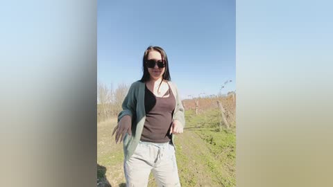 Video of a light-skinned woman with long brown hair, wearing sunglasses, a dark tank top, and light gray sweatpants, standing in a grassy, vineyard-like field under a clear blue sky.