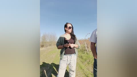 Video of a woman with long dark hair, wearing sunglasses, a green cardigan, and light gray pants, standing outdoors on a sunny day in a grassy field, holding a phone.