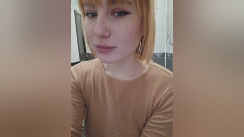 Video of a pale-skinned woman with short, straight, light-brown hair, wearing a long-sleeved, beige top, looking directly at the camera with a neutral expression. The background features white tiles and a blurred, dark object.