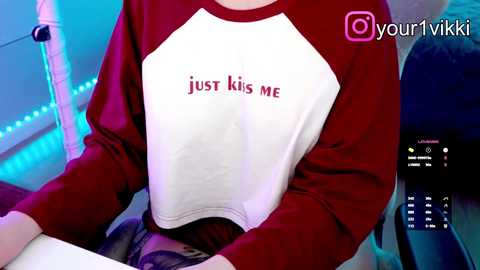 Video of a person wearing a white and maroon sweatshirt with \"just kiss me\" in red text. The background shows a blue-lit room with a gaming setup and a phone screen displaying a chat.