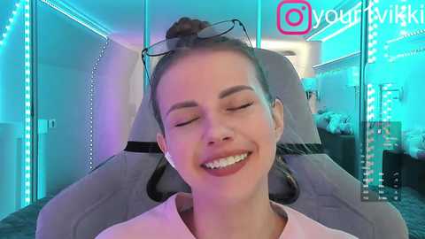 Video of a smiling, fair-skinned woman with brown hair in a bun, wearing glasses and a light pink top, seated in a grey gaming chair with blue LED lights. Background features a computer setup with social media icons.