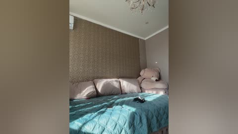 Video of a cozy, sunlit bedroom featuring a beige, patterned wallpaper backdrop, a plush beige couch, a teal quilted bedspread, a large beige teddy bear, and a crystal chandelier overhead.