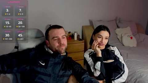 Video of a man in a dark jacket and woman in a black and white outfit sitting on a bed in a dimly lit room, with a virtual reality headset on the man.