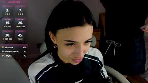 Video of a young woman with dark hair, wearing a white and black jacket, seated indoors. The background includes a dimly lit room with a wooden floor, a chair, and a person partially visible. A digital screen displays chat messages and a profile with a name \"Jenau.\