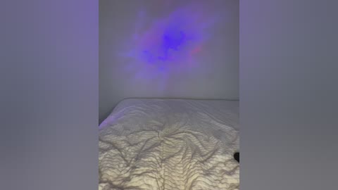 A video of a bed with a white, textured comforter featuring intricate, leaf-like patterns. The background is a plain, light gray wall with a faint, blue, nebula-like glow. The image conveys a serene, minimalist bedroom ambiance.
