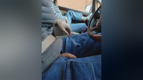 Video of a man in blue jeans and a gray sweater, leaning forward in a car, assisting an older man wearing a striped shirt and a brown jacket.