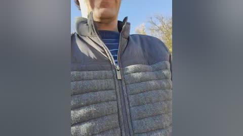 A close-up video of a person wearing a dark gray, quilted, zip-up jacket over a blue and white striped shirt. The background features a clear blue sky and autumn foliage. The image is centered on the chest and upper abdomen.