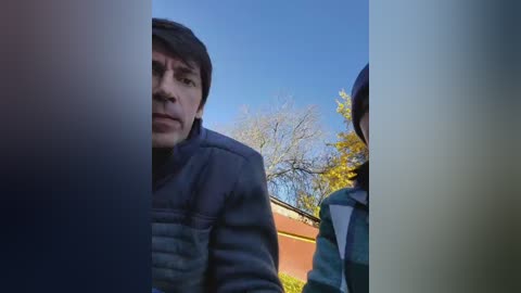 A video shows a man with short brown hair and a serious expression, wearing a dark jacket, standing in front of a car under a clear blue sky with autumn trees in the background.