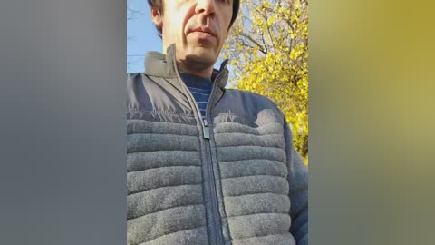 Video of a man wearing a gray, puffy jacket and a blue shirt, standing outdoors with yellow autumn leaves in the background.