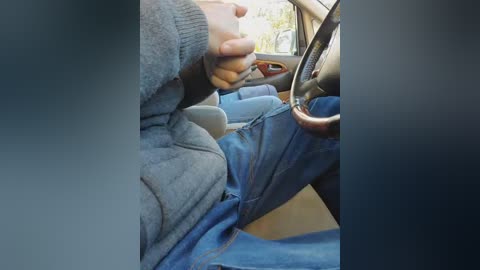 Video of a person wearing a grey sweater and blue jeans, seated in a car with their right hand gripping the steering wheel, and a blurred background of a vehicle interior.