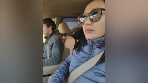 Video of a woman with light skin and straight black hair, wearing dark sunglasses and a blue puffer jacket, seated in a car, with another person in the background.