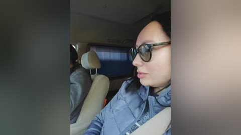 Video of a person with short dark hair and glasses, wearing a blue jacket, sitting in a car with beige seats and a blue sun visor.