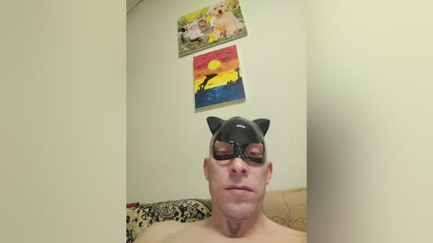 A video shows a shirtless, pale-skinned man with a black bat mask, reclining on a beige couch. Behind him, two framed paintings depict a sunset and a dog.