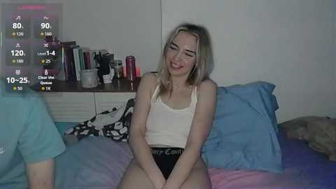 A video of a smiling young woman with light skin and blonde hair, wearing a white tank top and black panties, sitting on a bed with blue and purple sheets.