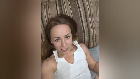 Video of a fair-skinned woman with long brown hair, wearing a white tank top, lying on a striped brown and beige couch, smiling.