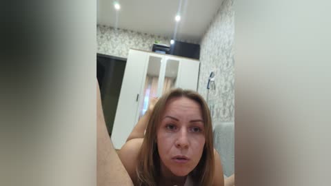 A video captures a Caucasian woman with long brown hair, wearing a white top, crouched in a hallway with white and patterned wallpaper. The angle is from a low perspective, emphasizing her facial expression.