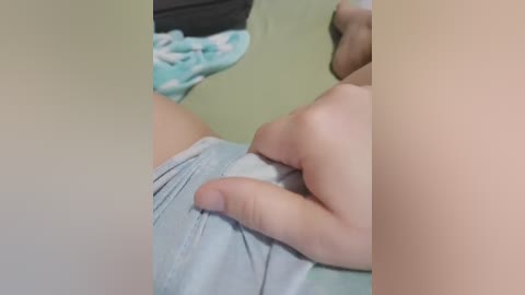 A close-up video of a person's hand adjusting light blue shorts, showing a light-skinned arm and a blurred background with green and white patterned fabric.