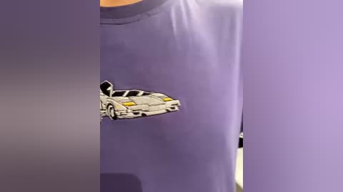 Video of a person wearing a light purple T-shirt with a detailed, cartoon-style drawing of a white sports car on the front. The car has yellow headlights and a raised hood, with the image slightly blurred. The background is a soft purple hue.
