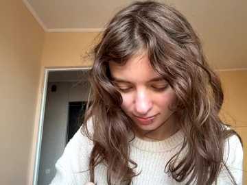Video of a young woman with long, wavy brown hair, wearing a white sweater, standing indoors in a beige room with a doorway in the background.
