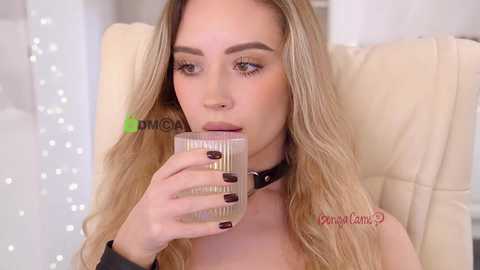 Video of a fair-skinned woman with long blonde hair, wearing a black choker, sitting in a cream-colored chair, drinking from a glass, nails painted dark, blurred background.