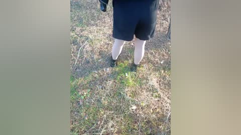 Video of a person's legs in black shorts and white socks, standing on a dry, grassy ground with sparse patches of green, surrounded by dry, brown grass and twigs.